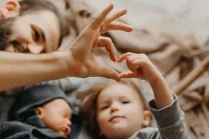A man and his two children are lying on a bed and are making a heart shape with their hands. The child on the right is wearing a hat, while the child on the left is sleeping. The man is looking at them.