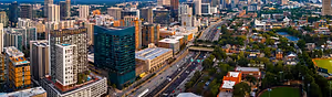 An aerial view of a city with many buildings and skyscrapers, including a tall building in the distance, surrounded by trees, with a wide road, cars, and a sunset in the distance