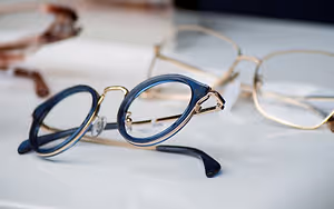 Three pairs of glasses, one with a blue frame, one with a gold frame, and one with a brown frame, are displayed on a white surface, with the blue-framed glasses positioned in the front, and the gold-framed glasses in the back.