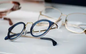 Three pairs of glasses, one with a blue frame, one with a gold frame, and one with a brown frame, are displayed on a white surface, with the blue-framed glasses positioned in the front, and the gold-framed glasses in the back.