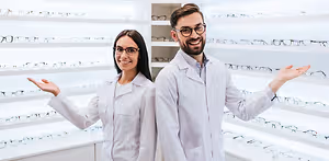 A smiling man and woman wearing white lab coats and glasses standing next to each other in front of a wall with rows of eyeglasses on display