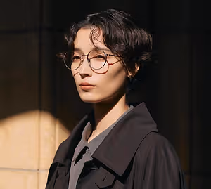 Asian woman with short hair wearing glasses and a black coat posing in front of a building