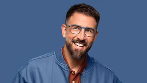 A smiling man with a beard, wearing glasses and a blue jacket, posing for a photo