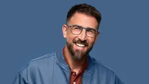 A smiling man with a beard, wearing glasses and a blue jacket, posing for a photo