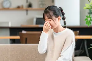 A young girl is sitting on a couch and blowing her nose with a tissue in a living room.