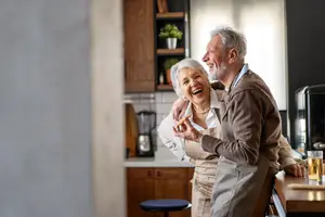 An elderly man and woman stand in a kitchen, the woman laughing and the man smiling as he looks at her and holds food.