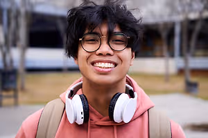 A young man wearing glasses, a pink hoodie, and white headphones with a backpack on his back, smiling and posing for a photo in an outdoor area