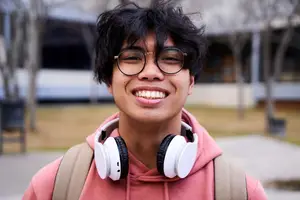 A young man wearing glasses, a pink hoodie, and white headphones with a backpack on his back, smiling and posing for a photo in an outdoor area