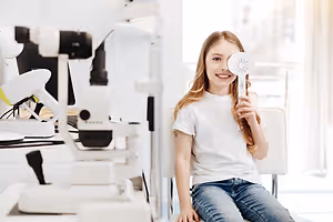 A smiling girl is sitting on a chair in a room with medical equipment and a large window, holding a white fan.