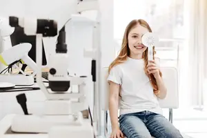 A smiling girl is sitting on a chair in a room with medical equipment and a large window, holding a white fan.
