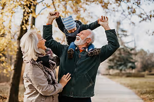 An elderly man lifts a child up and a woman stands beside him in a park with trees in autumn