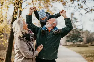 An elderly man lifts a child up and a woman stands beside him in a park with trees in autumn