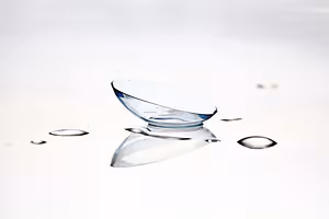 Close-up of a clear glass contact lens sitting on a smooth white surface with a few water droplets around it.