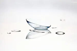 Close-up of a clear glass contact lens sitting on a smooth white surface with a few water droplets around it.