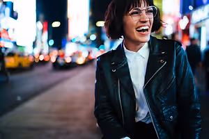A smiling woman wearing glasses and a black leather jacket on a city street at night.