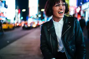 A smiling woman wearing glasses and a black leather jacket on a city street at night.