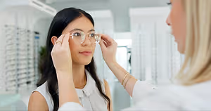 A woman is adjusting glasses on another woman in a white room