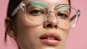 A woman with glasses and freckles looks at the camera against a pink background
