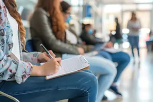 A woman in a hospital or clinic waiting room, holding a clipboard and pen, with other people sitting and standing behind her.