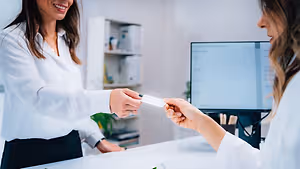 Two women in white lab coats are standing at a counter, one is handing a card to the other