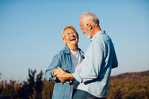 An old man and woman are standing in a field, smiling and looking at each other.