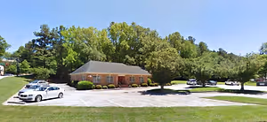 A house in a parking lot with cars parked in front of it and trees in the background