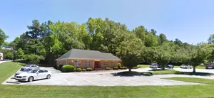 A house in a parking lot with cars parked in front of it and trees in the background