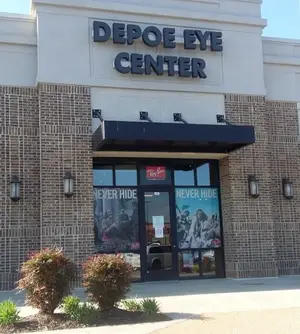 The exterior of the Depoe Eye Center with a brick wall, glass doors, and a black awning with the text 'Depoe Eye Center' and 'Never Hide' on the glass. There are plants and lamps on either side of the entrance.