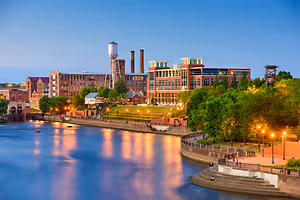 A city scene at dusk with a river, buildings, trees, and streetlights.
