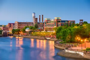A city scene at dusk with a river, buildings, trees, and streetlights.