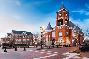 A town square with a clock tower, benches, and street lights in a brick pavement area under a blue sky