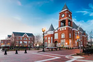 A town square with a clock tower, benches, and street lights in a brick pavement area under a blue sky