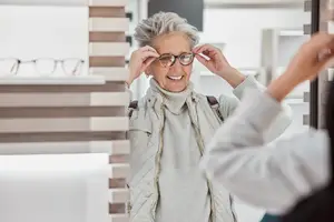 A smiling woman with gray hair is trying on glasses in front of a mirror at a store