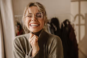 A woman with glasses is smiling in a clothing store with a white curtain and clothes on hangers behind her