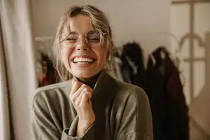 A woman with glasses is smiling in a clothing store with a white curtain and clothes on hangers behind her
