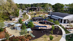 Aerial view of a park with various structures, including a large play area, gazebos, and a black roof, surrounded by trees and greenery.