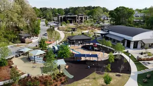 Aerial view of a park with various structures, including a large play area, gazebos, and a black roof, surrounded by trees and greenery.