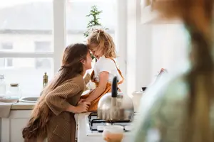 A woman and a young girl are in the kitchen, the woman is holding the girl while the girl is holding a bowl of food and smiling, the kitchen has a stove with a kettle and a window with a view of the outside.