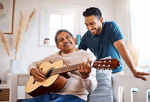 A man in a blue t-shirt is smiling as he watches an older man in a white sweater playing the guitar.