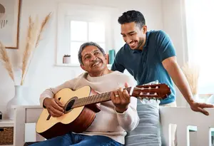 A man in a blue t-shirt is smiling as he watches an older man in a white sweater playing the guitar.