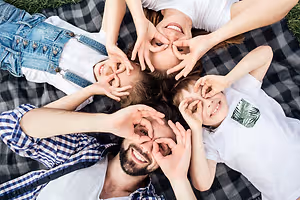 A family of four, two adults and two kids, are laying on a blanket and making a heart shape with their hands.