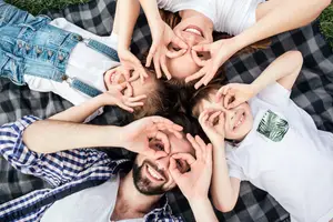 A family of four, two adults and two kids, are laying on a blanket and making a heart shape with their hands.