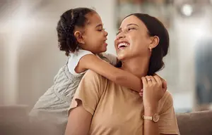 A woman is smiling while holding a girl child on her shoulder inside a room, probably in their living room.