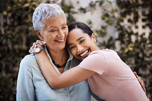 An older woman with white hair wearing a blue shirt and a necklace hugging a young woman wearing a pink shirt and smiling in a garden with a white wall in the background.