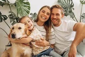 A family of three is sitting on a couch, smiling and posing for a photo with a dog in front of them, and they are all wearing casual clothing