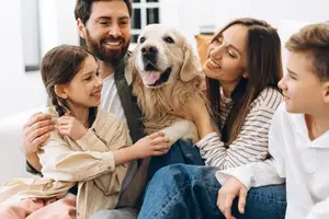A family, including two adults, two kids, and a golden retriever, is sitting on a couch, smiling and looking at the camera. Behind them, a white wall with a window is visible.