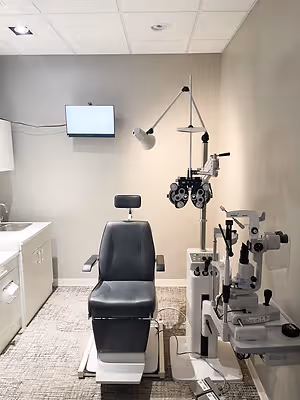 An interior view of an eye clinic room featuring a patient chair, ophthalmoscope, and a monitor mounted on the wall.