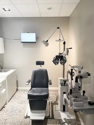 An interior view of an eye clinic room featuring a patient chair, ophthalmoscope, and a monitor mounted on the wall.