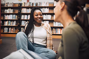 A smiling girl with braided hair is sitting on the floor with her legs crossed in the library while talking to another girl who is holding a book