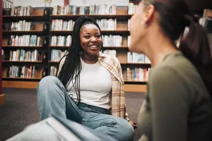 A smiling girl with braided hair is sitting on the floor with her legs crossed in the library while talking to another girl who is holding a book
